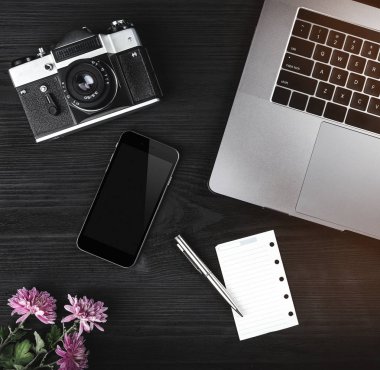 Siyah ahşap zemin üzerine notebook klavye, kamera, cep telefonu, Gözlük, Sevimli çiçek ve notlar sayfasını çalışma alanının üstten görünüm