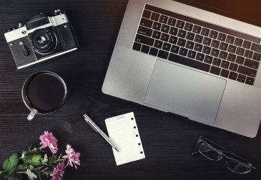 Bir fincan çay veya kahve, fotoğraf makinesi, laptop bilgisayar, gözlük ve pembe çiçekler siyah ahşap masa üzerinde. Üstten görünüm, düz yatıyordu