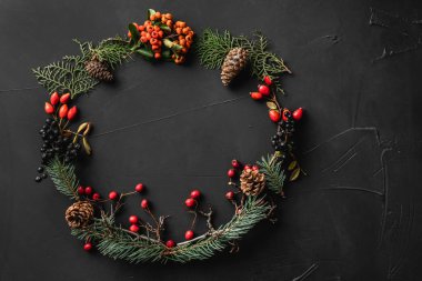 Crown of Christmas tree branches, pine cones, berries, nuts, on black stone background. Xmas and Happy New Year theme. Flat lay, top view clipart