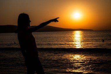 Sahilde yaz güneşinin doğuşunu ya da günbatımını seyreden bir kız siluetinin arka görüntüsü.