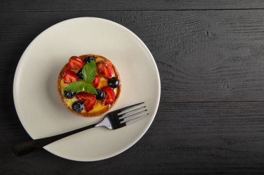 Siyah çatallı beyaz bir tabakta meyveli tart veya muhallebi, siyah ahşap arka planda kopyalama alanı, üst manzara