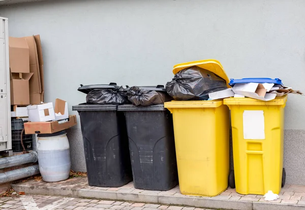 Overflowing plastic waste cans with black garbage bags, cardboard boxes and a container with a liquid