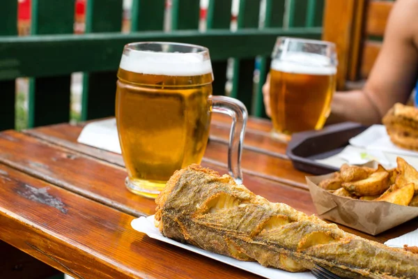 Delicious fried fish and potatoes on disposable paper plate and chilled glass of draft beer on wooden table in outdoor cafe. Selective focus — Stok fotoğraf