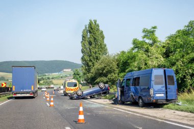Otoyolda araba kazası. Çarpışmadan sonra mavi minibüs hasar görmüş, yolda araba ve ambulans devrilmiş. Trafik konileri kaza mahallinde