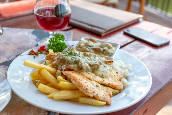 Beyaz mantar soslu ızgara tavuk filetosu ve beyaz tabakta patates kızartması. Arka planda bir kadeh kırmızı şarap.