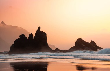 Manzara ve dalgalar Tenerife 'deki Benijo' nun siyah kumsalında güzel turuncu bir günbatımında kayalara çarpıyor.