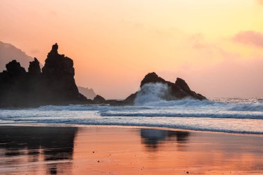 Manzara ve dalgalar Tenerife 'deki Benijo' nun siyah kumsalında güzel turuncu bir günbatımında kayalara çarpıyor.
