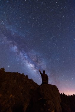 Geniş kollu genç adam dağlarda bir yaz gecesinde yıldızları ve samanyolu işaret ediyor.