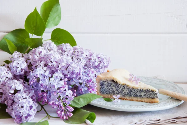 Slice of poppy seed cake with lilac flowers on white wooden background