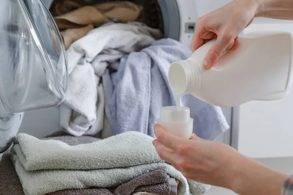 Primer plano de las manos femeninas vertiendo detergente líquido de lavandería en la tapa. Lavadora y ropa con cesta de mimbre en el fondo — Foto de Stock