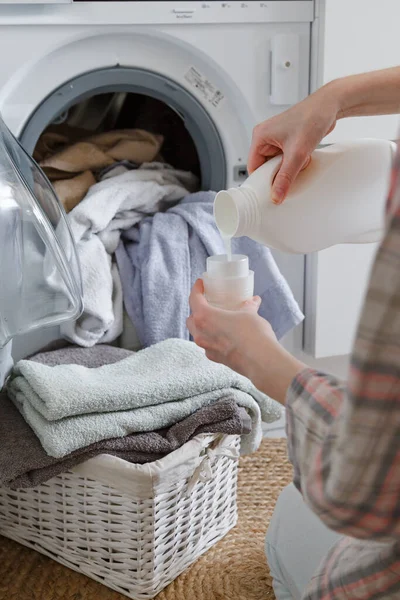 Primer plano de las manos femeninas vertiendo detergente líquido de lavandería en la tapa. Lavadora y ropa con cesta de mimbre en el fondo — Foto de Stock