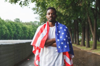 Omzunda Amerikan bayrağı olan yakışıklı Afro-Amerikan bir adam ciddi ciddi kameraya bakıyor, dışarıda duruyor. Yaz günü