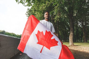 Handsome Afro American man with Canadian flag on his shoulders seriously looking at camera, standing outdoors. Day, summer clipart