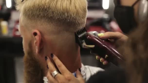 Egy női borbély maszkban divatos frizurát csinál egy szakállas fiatalembernek hajvágóval. A borbélyüzlet a világjárvány után is nyitva tart. — Stock videók
