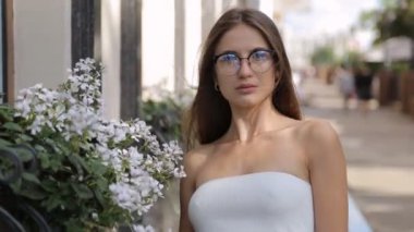 Uzun saçlı, gözlüklü, caddedeki çiçeklerin yanında duran çekici bir genç kadın.