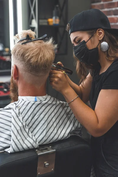 Female barber in mask cuts a man hairs with hair clipper. Hairstyle during social distancing.