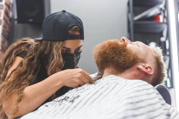 A man at a barbershop. Woman barber clipping beard and mustache. Barber woman in mask.