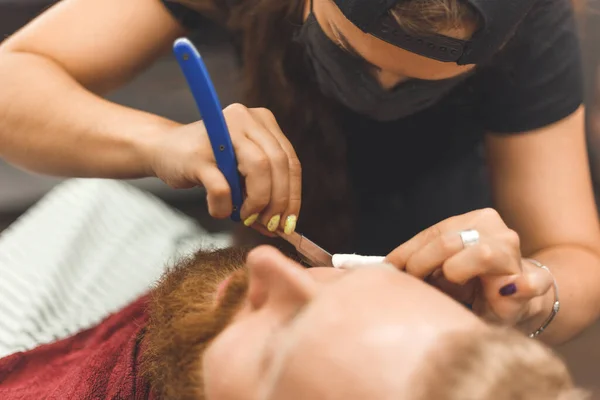 Barber woman shaves beard with a straight razor. Hairdresser equipment. Selective focus. Master in face mask.