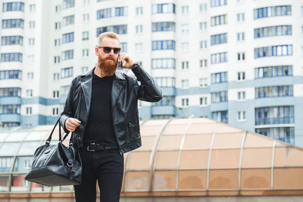 Fashionably dressed bearded caucasian man walks around the city with leather bag and speaking on the phone. Urban casual style