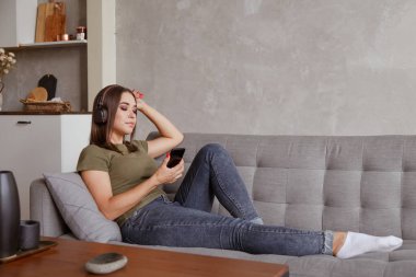 Genç esmer kadın kablosuz kulaklıklarla müzik dinliyor akıllı telefona bağlı, evde tek başına kanepede yatıyor.