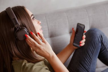 Genç esmer kadın kablosuz kulaklıklarla müzik dinliyor akıllı telefona bağlı, evde tek başına kanepede yatıyor.