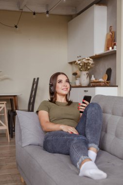 Genç esmer kadın kablosuz kulaklıklarla müzik dinliyor akıllı telefona bağlı, evde tek başına kanepede yatıyor.