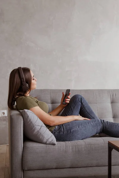 Junge brünette Frau genießt die Musik in drahtlosen Kopfhörern, die mit dem Smartphone verbunden sind, allein zu Hause auf dem Sofa liegend — Stockfoto