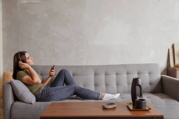 Joven morena disfrutando de la música en auriculares inalámbricos conectados a smartphone, tumbada en el sofá en casa sola — Foto de Stock