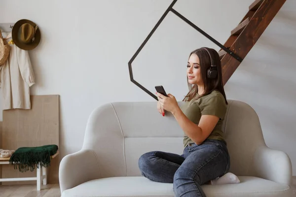 Jovencita alegre escuchando música en auriculares inalámbricos conectados a smartphone en apartamento loft — Foto de Stock