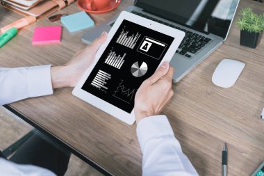 İşadamı ahşap masadaki dijital tablet, kitap, belge ve dizüstü bilgisayarla çalışıyor. Finansal veri içeren tablet ekranı.