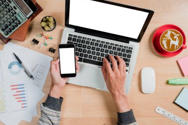 Ofiste boş ekran dizüstü bilgisayar, akıllı telefon ve çalışma alanı üzerinde çalışan bir adam. Ürün gösterimi için boş ekranlı üst görünüm.