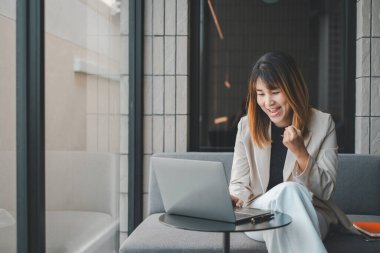 Bilgisayarlı neşeli genç iş kadını modern ofis yerinde oturmuş başarılı bir anı kutluyor..