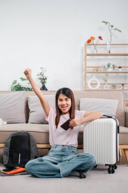 Bavul ve pasaportla yerde oturan mutlu gezgin şık bir oturma odasında seyahat planlarını kutluyor..
