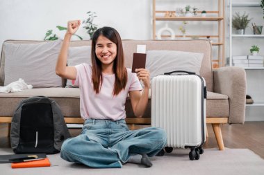 Pasaportu ve bavuluyla oturan gülümseyen genç bayan, seyahat etmeye hazır. Konforlu oturma odası arka planı.