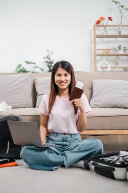 Gülümseyen kadın dizüstü bilgisayarı ve pasaportuyla yerde oturuyor, seyahat etmeye hazır. Konforlu oturma odası arka planı.