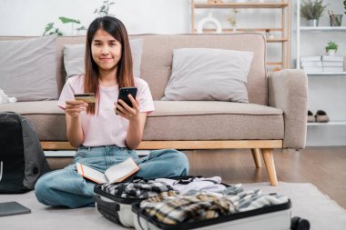Kadın yerde oturmuş bavulunu topluyor, akıllı telefon ve kredi kartını kullanarak rahat bir oturma odasında online alışveriş yapıyor..