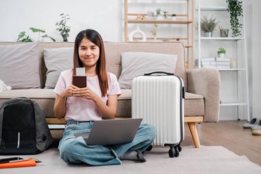 Gülümseyen kadın seyahate hazırlanıyor, elinde laptop ve bavuluyla modern bir oturma odasında oturuyor..