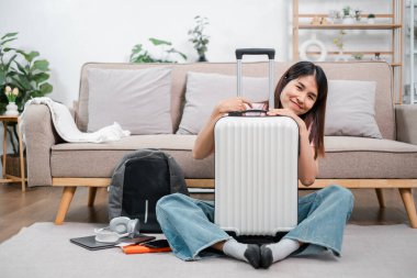 Gülümseyen kadın bavuluyla yerde oturuyor, seyahat etmeye hazır. Modern oturma odası, günlük kıyafetler ve seyahat gereksinimleri..