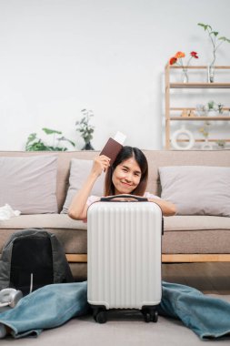 Elinde bavul ve pasaportuyla gülümseyen bir kadın, rahat bir oturma odasında yerde oturuyor, seyahat etmeye hazır..