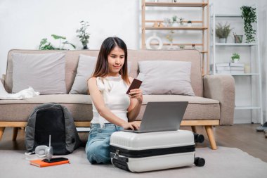 İnternetten alışveriş yapmak için dizüstü bilgisayar ve kredi kartı kullanan bir kadın modern bir oturma odasında bavulun üzerinde oturuyor..
