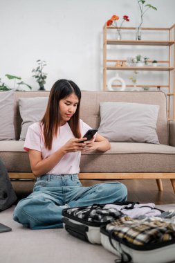 Genç bir kadın yerde oturmuş bavulunu topluyor, şık bir oturma odasında akıllı telefonunu kullanıyor, geziye hazırlanıyor..