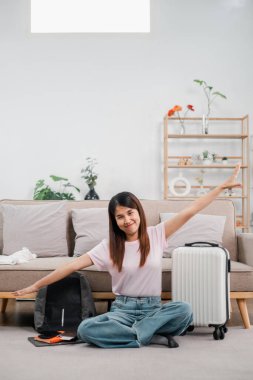 Sırt çantası ve bavuluyla yerde oturan gülümseyen kadın, seyahat etmeye hazır. Bitkileri ve dekorları olan modern oturma odası..