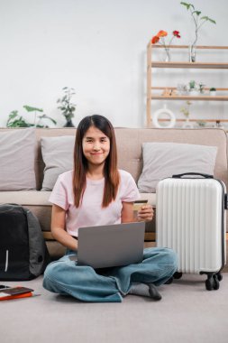 Yerde dizüstü bilgisayarı ve kredi kartıyla oturan bir kadın, seyahat etmeye hazır. Bitkiler ve bavullarla dolu rahat bir ev..