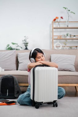 Kulaklık takan genç bir kız, rahat bir oturma odasında, seyahat etmeye hazır bir bavulun üzerinde dinleniyor..