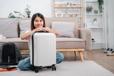 Oturma odasında bavuluyla yerde oturan gülümseyen kadın, seyahat etmeye hazır. Bitkiler ve raflarla modern iç mekan.