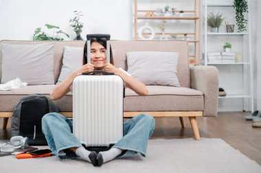 Oturma odasında bavuluyla oturan gülümseyen kadın, seyahat etmeye hazır. Modern dekorasyon ve seyahat ögeleri.