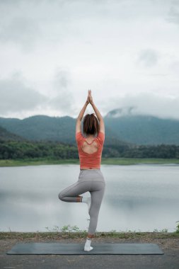 Bir kadın, bulutlu bir gökyüzünün altında, arkasında dağlar olan sakin bir gölün yanında yoga ağacı pozu veriyor..