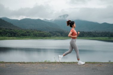 Arka planda dağlar olan göl kenarında koşan bir kadın spor salonu ve açık hava yaşam tarzı sergiliyor..