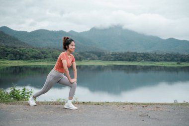 Aktif kıyafetli bir kadın gölün kenarında geriniyor. Arka planda dağlar var. Spor salonu ve doğa..