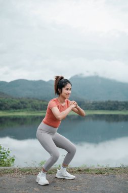 Aktif giysili bir kadın gölün kenarında çömelmiş, arka planda dağlar, açık hava fitness ve doğa gösterileri yapıyor..